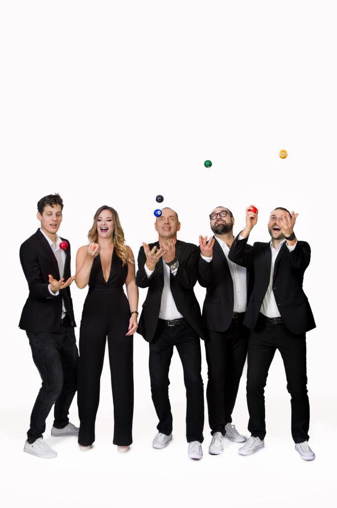 Pięcioosobowa grupa w czarnych strojach, w tym jedna kobieta w kombinezonie, żongluje kolorowymi piłkami na białym tle.