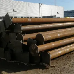 Stalowe słupy z przyspawanymi płytami bazowymi, ułożone w stos na placu budowy, widoczne ślady korozji, w tle budynek przemysłowy.