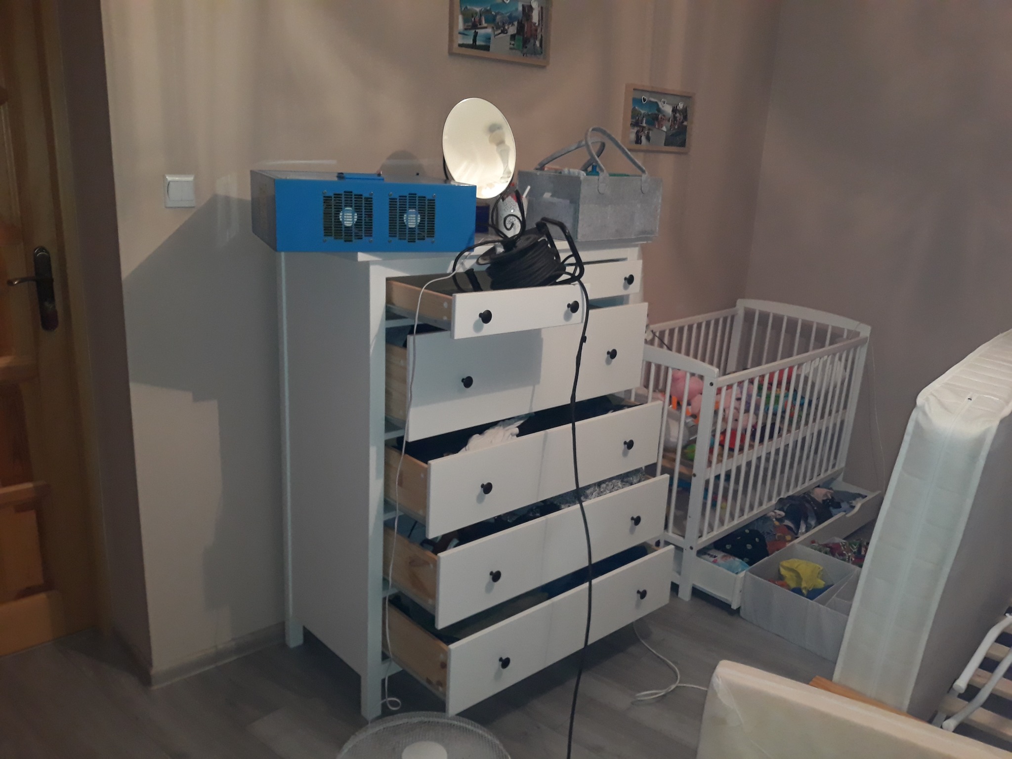 Niebieski osuszacz powietrza na białej komodzie z otwartymi szufladami, obok dziecięcego łóżeczka i materaca.