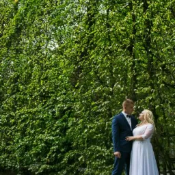 Para młoda w eleganckich strojach stoi na tle gęstego, zielonego żywopłotu w parku, patrząc na siebie z uśmiechem.