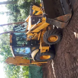 Żółta koparko-ładowarka JCB 3CX w trakcie prac ziemnych, widoczne ślady opon na ubitej ziemi, drzewa w tle.