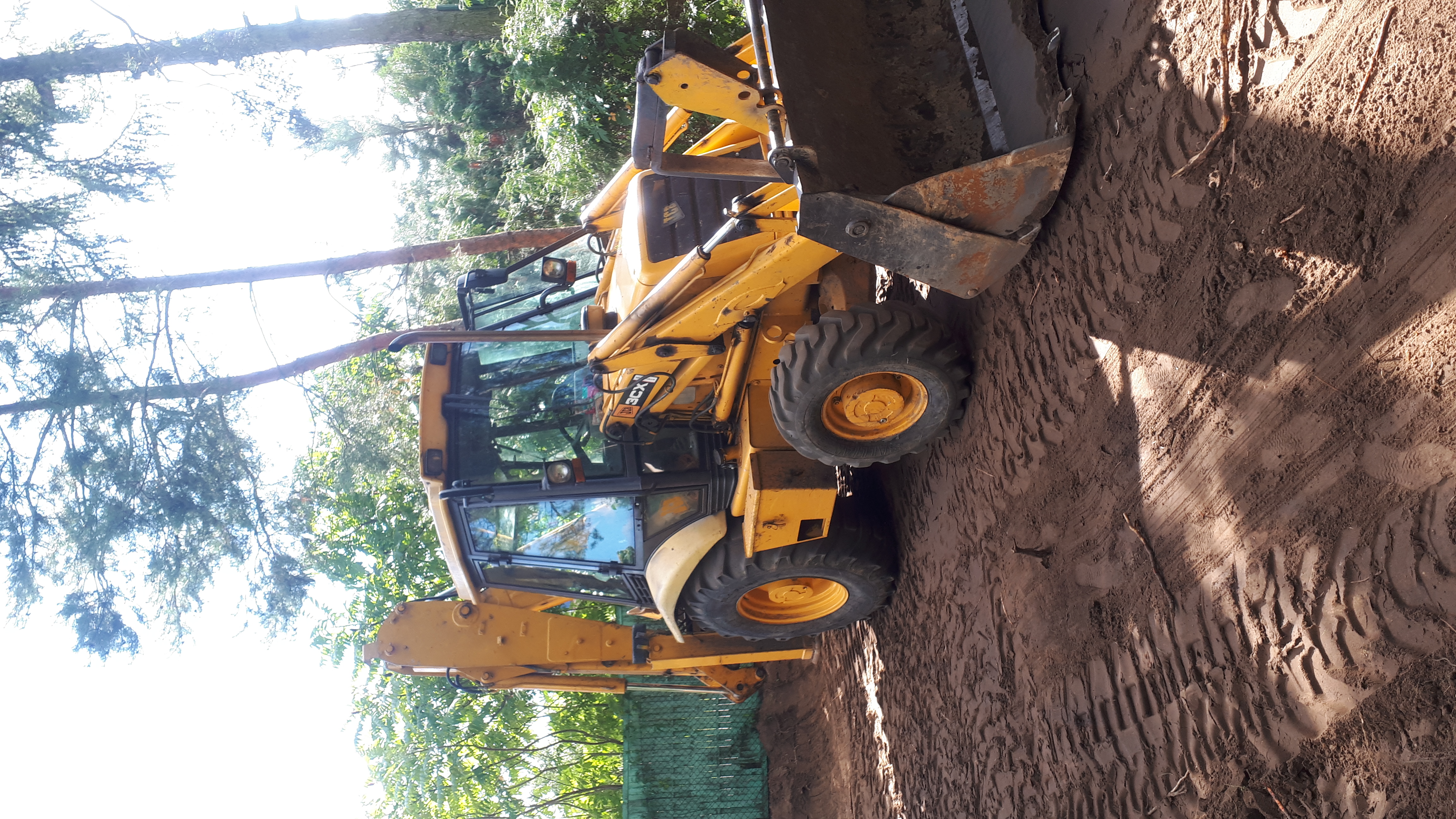 Żółta koparko-ładowarka JCB 3CX w trakcie prac ziemnych, widoczne ślady opon na ubitej ziemi, drzewa w tle.