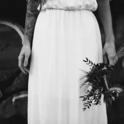 Czarno-białe zdjęcie panny młodej w białej sukni, trzymającej bukiet polnych kwiatów i piór, z tatuażem na ramieniu i graffiti w tle.