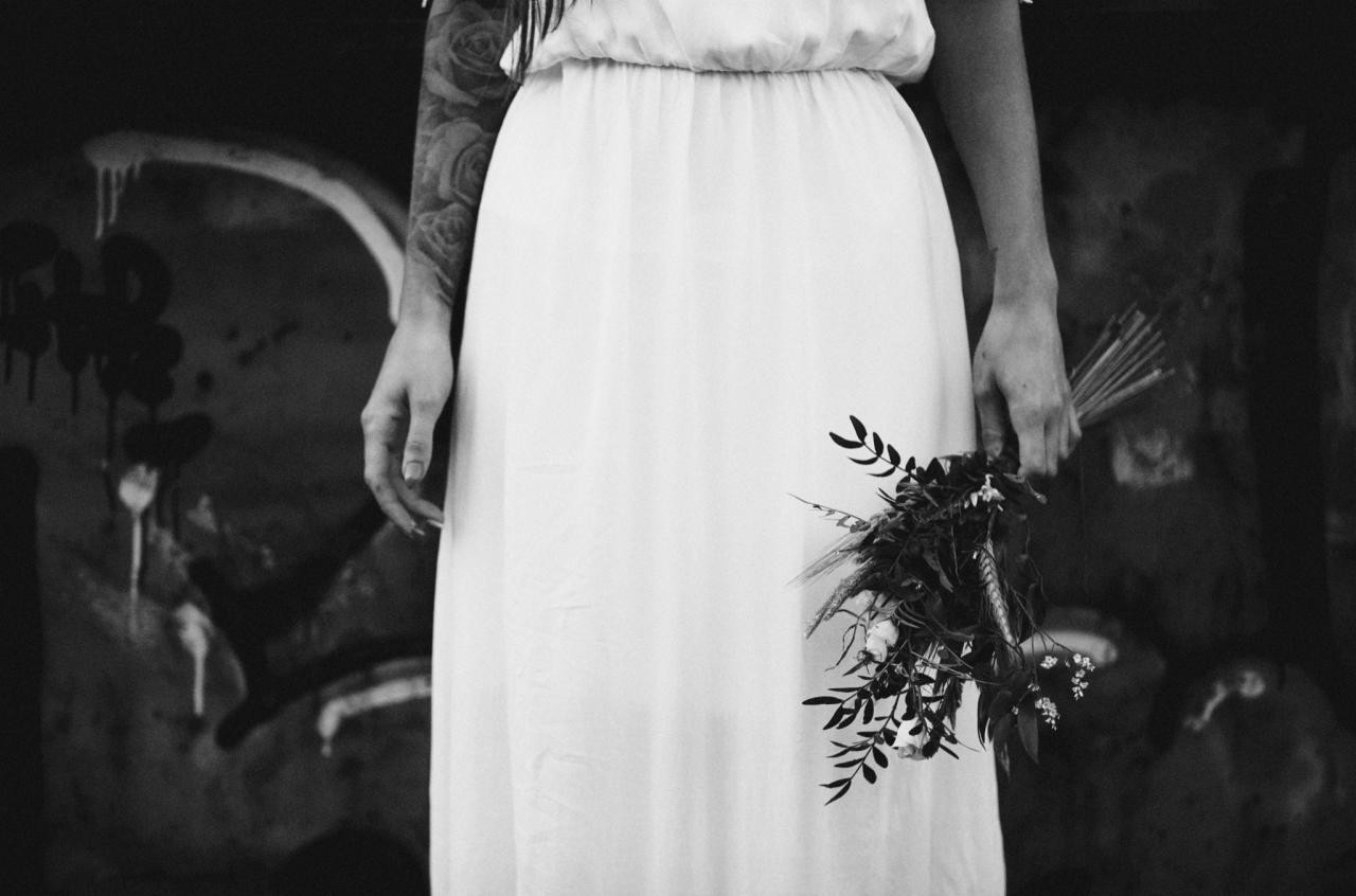 Czarno-białe zdjęcie panny młodej w białej sukni, trzymającej bukiet polnych kwiatów i piór, z tatuażem na ramieniu i graffiti w tle.