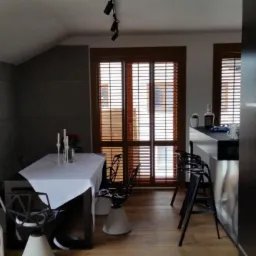 Jasne wnętrze pokoju z szarymi panelami ściennymi, stołem nakrytym białym obrusem, nowoczesnymi krzesłami i drewnianymi żaluzjami w oknach.