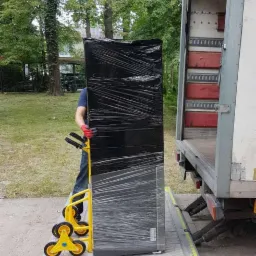 Lodówka owinięta folią stretch na wózku schodowym z żółtymi kołami, przygotowywana do załadunku na platformę samochodu dostawczego na tle zieleni.