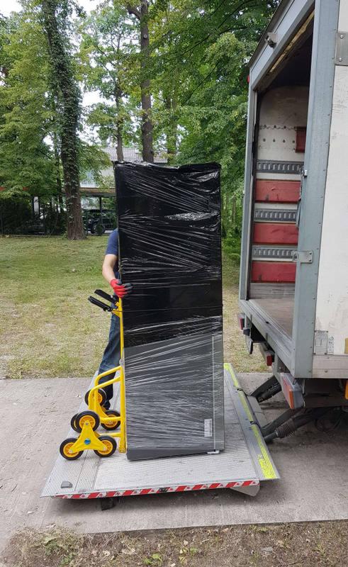 Lodówka owinięta folią stretch na wózku schodowym z żółtymi kołami, przygotowywana do załadunku na platformę samochodu dostawczego na tle zieleni.