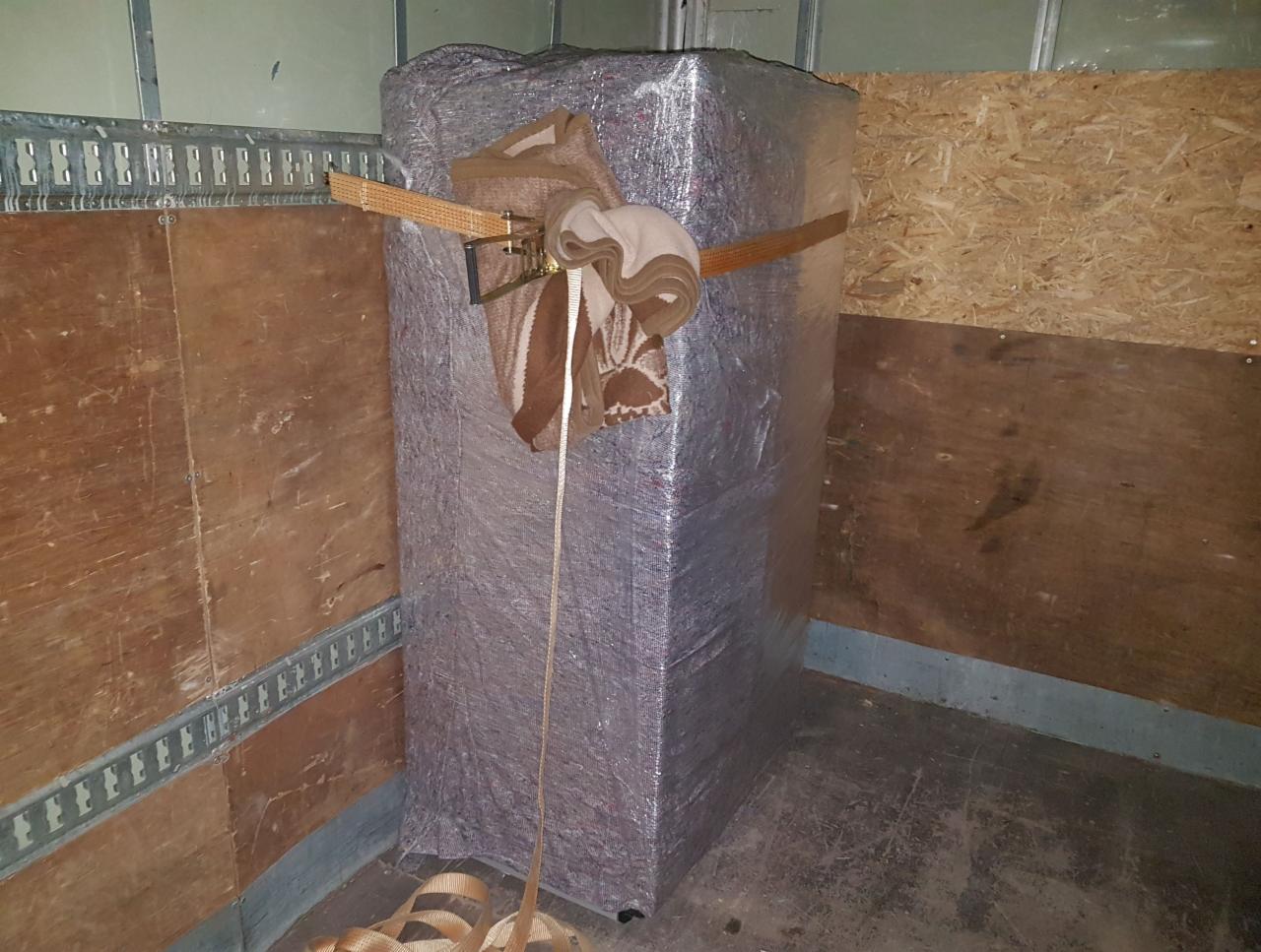 Zabezpieczony mebel w folii i pasach transportowych wewnątrz przestrzeni ładunkowej, widoczne drewniane ściany i podłoga pojazdu.