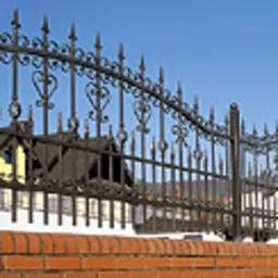 Kute, metalowe ogrodzenie z dekoracyjnymi elementami w kształcie serc i grotów, osadzone na niskim, ceglanym murku. W tle widoczny fragment dachu domu.