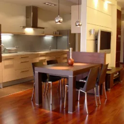 Nowoczesna jadalnia z drewnianym stołem, czarnymi krzesłami i parkietem w odcieniach brązu, otwarta na kuchnię z zabudową w jasnym drewnie i srebrnymi akcentami, widoczny fragment salonu...