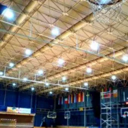 Wnętrze hali sportowej oświetlone szeregiem lamp zamontowanych pod metalową konstrukcją dachu, widoczne kosze do koszykówki i flagi państw.
