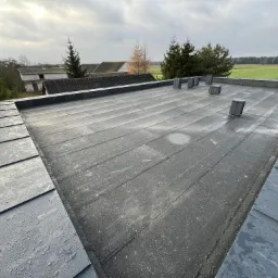 Płaski dach pokryty papą z metalowymi wywietrznikami, widok z góry na tle krajobrazu z polami i zabudowaniami w pochmurny dzień.