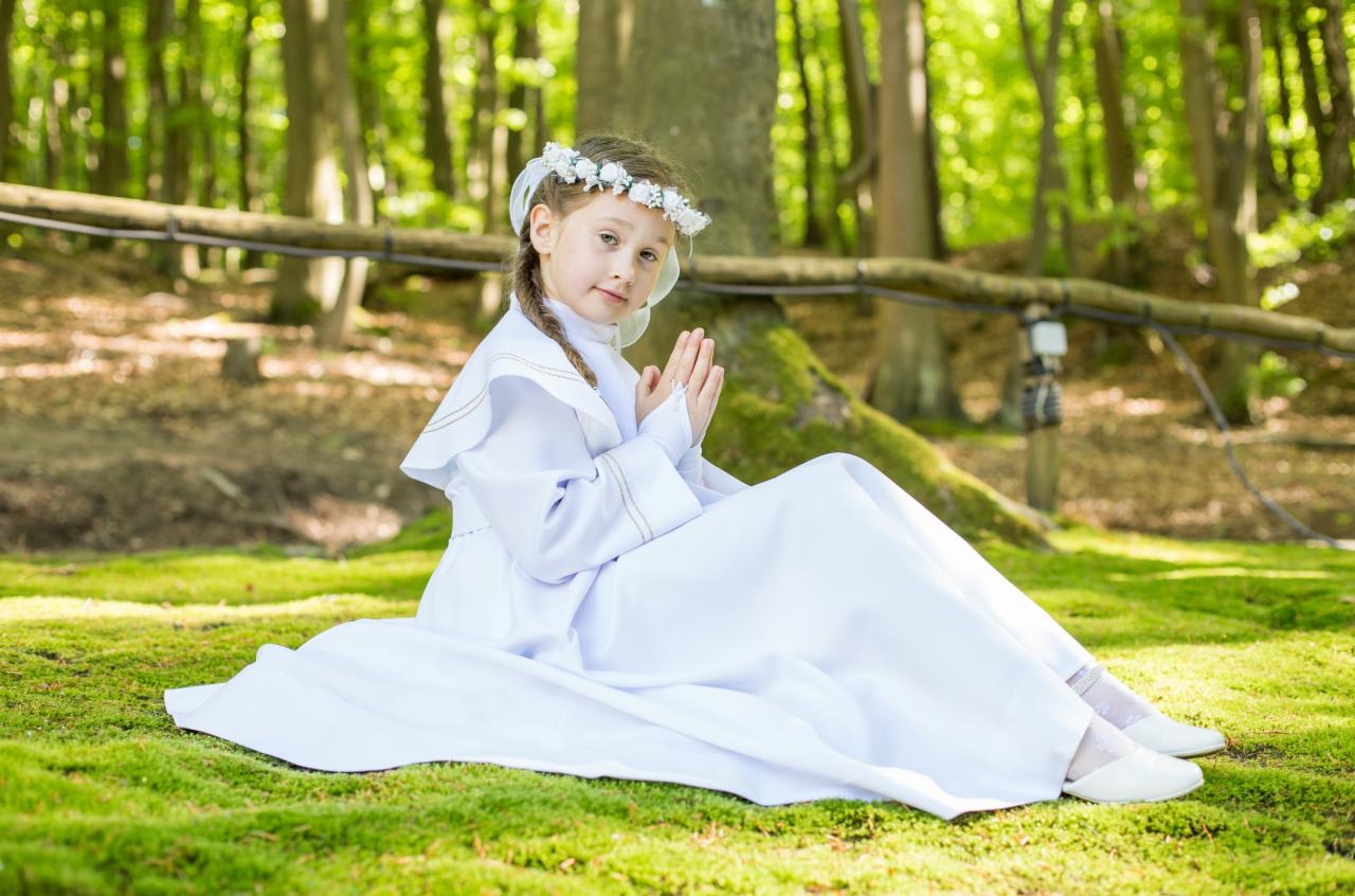 Dziewczynka w białej albie i wianku na głowie siedzi na trawie w lesie, z rękami złożonymi do modlitwy.