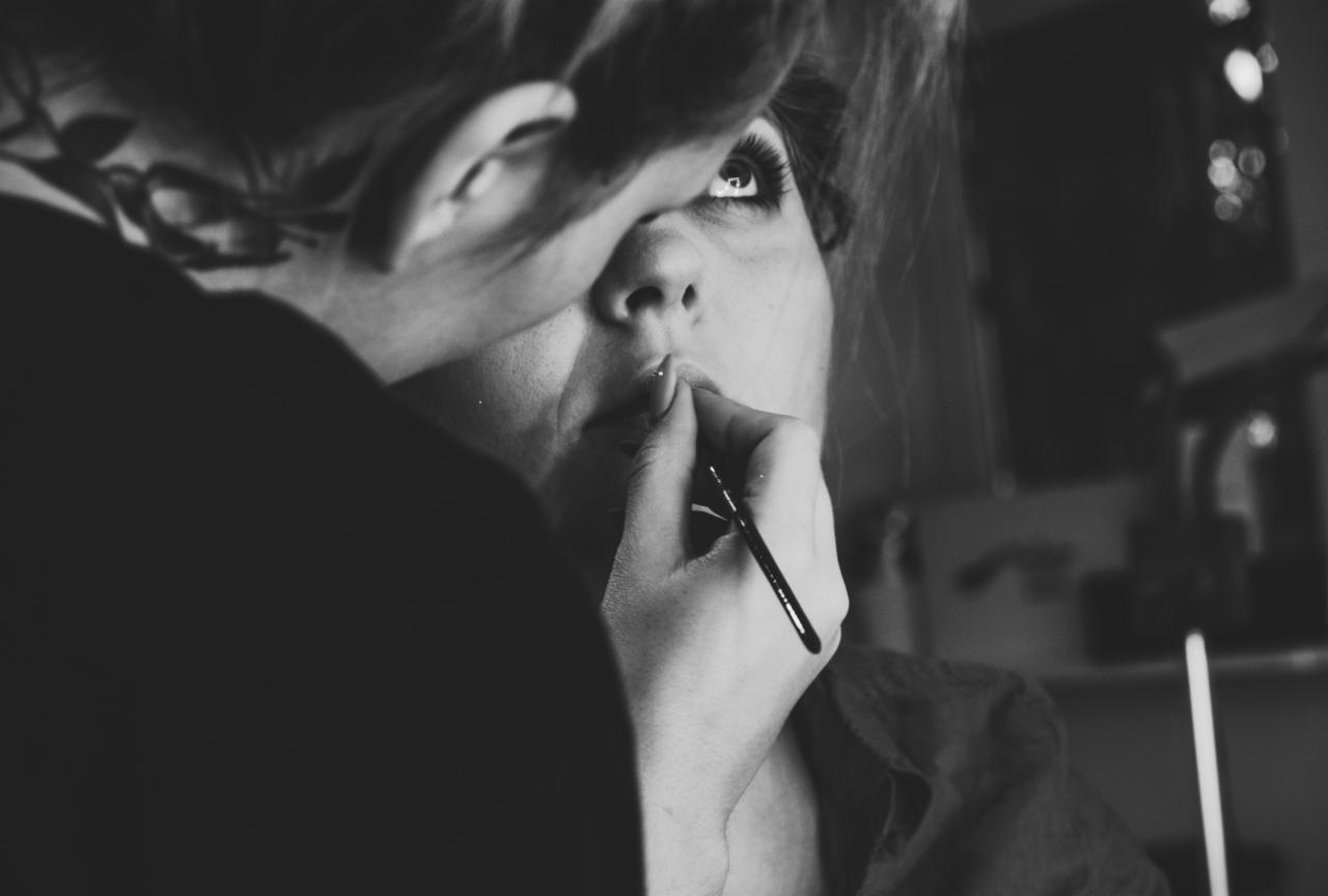Czarno-białe ujęcie kobiety podczas nakładania makijażu na usta pędzelkiem, z widocznymi długimi rzęsami i tatuażem za uchem.