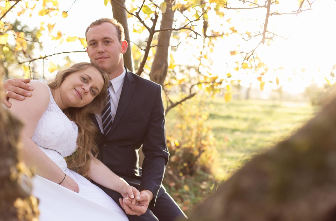 Para młoda w jesiennej scenerii siedzi przy drzewie, panna młoda w białej sukni z koronkową górą opiera głowę o ramię pana młodego w ciemnym garniturze, delikatne światło słoneczne przebija przez...