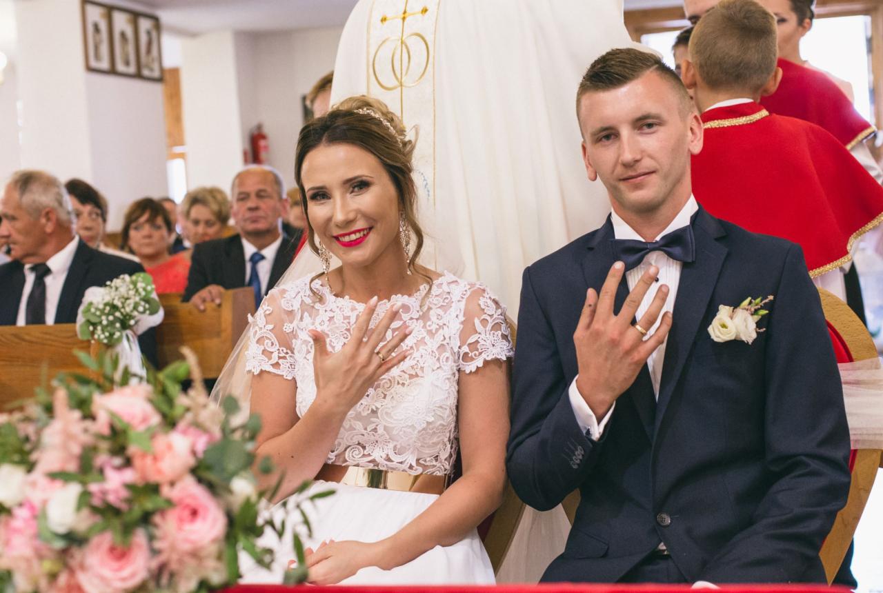 Para młoda w kościele pokazuje obrączki, panna młoda w koronkowej sukni z szerokim złotym pasem, pan młody w granatowym garniturze z muchą, w tle goście i kwiaty.