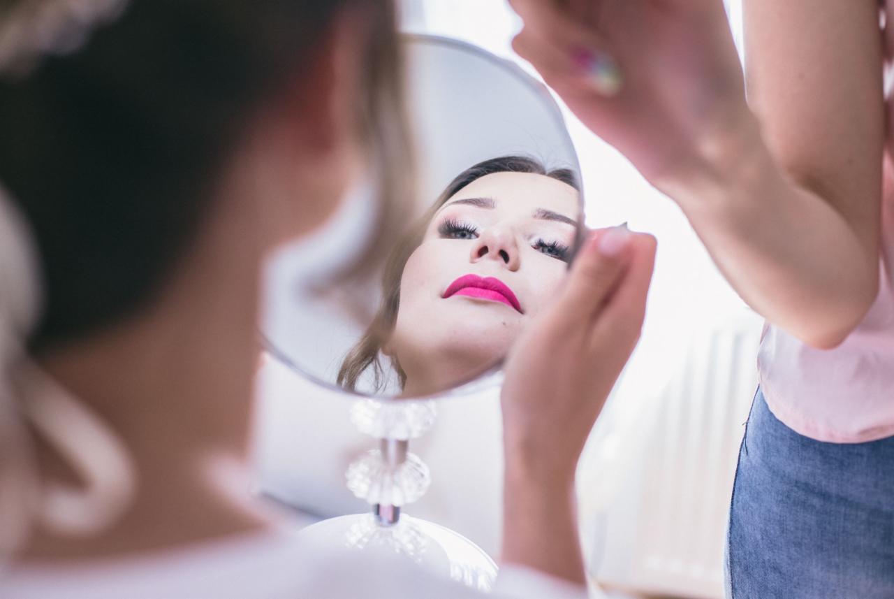 Kobieta z intensywnym makijażem ust odbija się w okrągłym lustrze na ozdobnej nóżce, w tle widoczna dłoń w trakcie wykonywania makijażu.