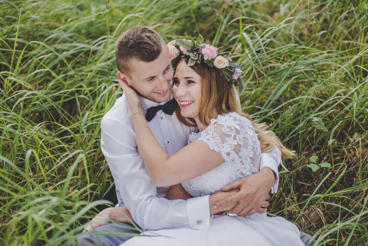 Uśmiechnięta para młoda w objęciach, siedząca w wysokiej trawie, panna młoda z wiankiem z róż na głowie, pan młody w muszce.