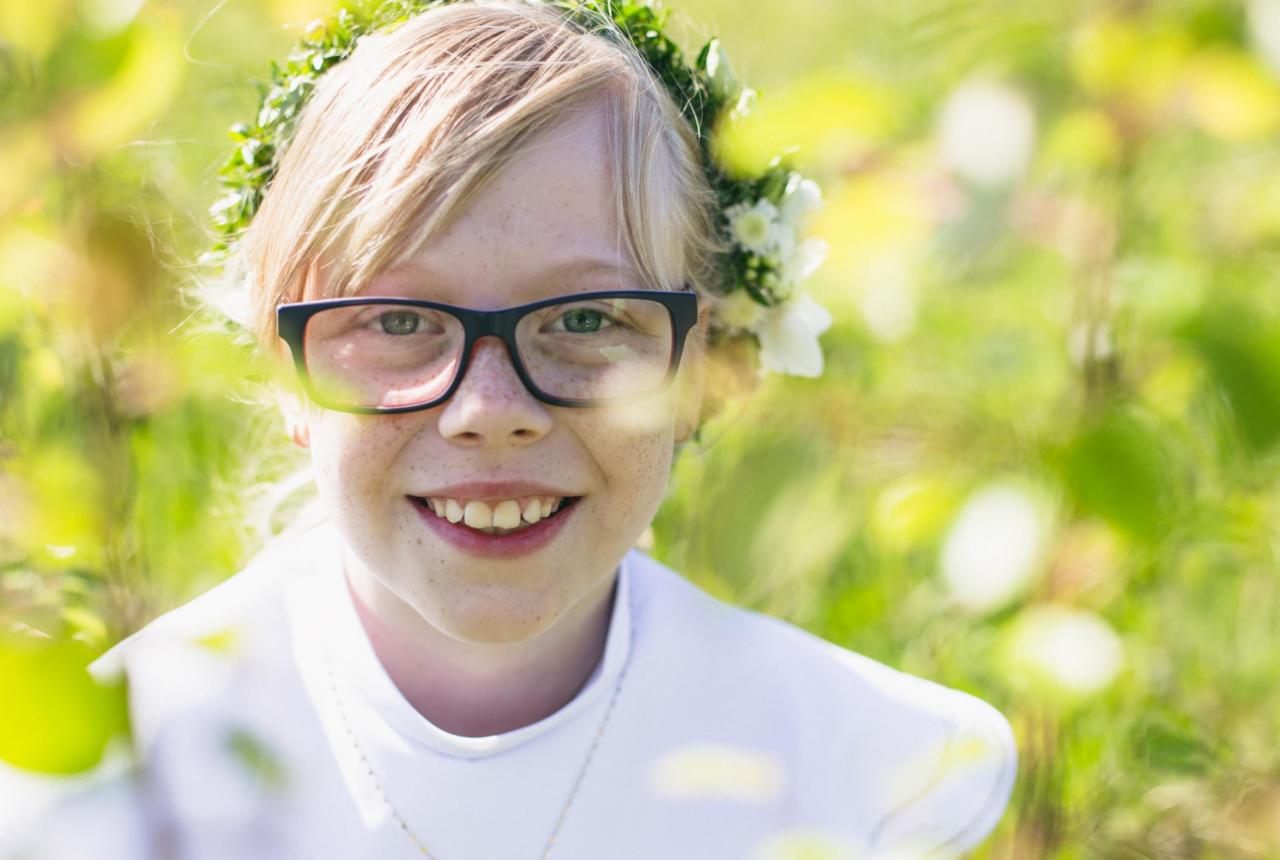 Uśmiechnięta dziewczynka w okularach i wianku z białych kwiatów na głowie, ubrana w białą albę, na tle zieleni.