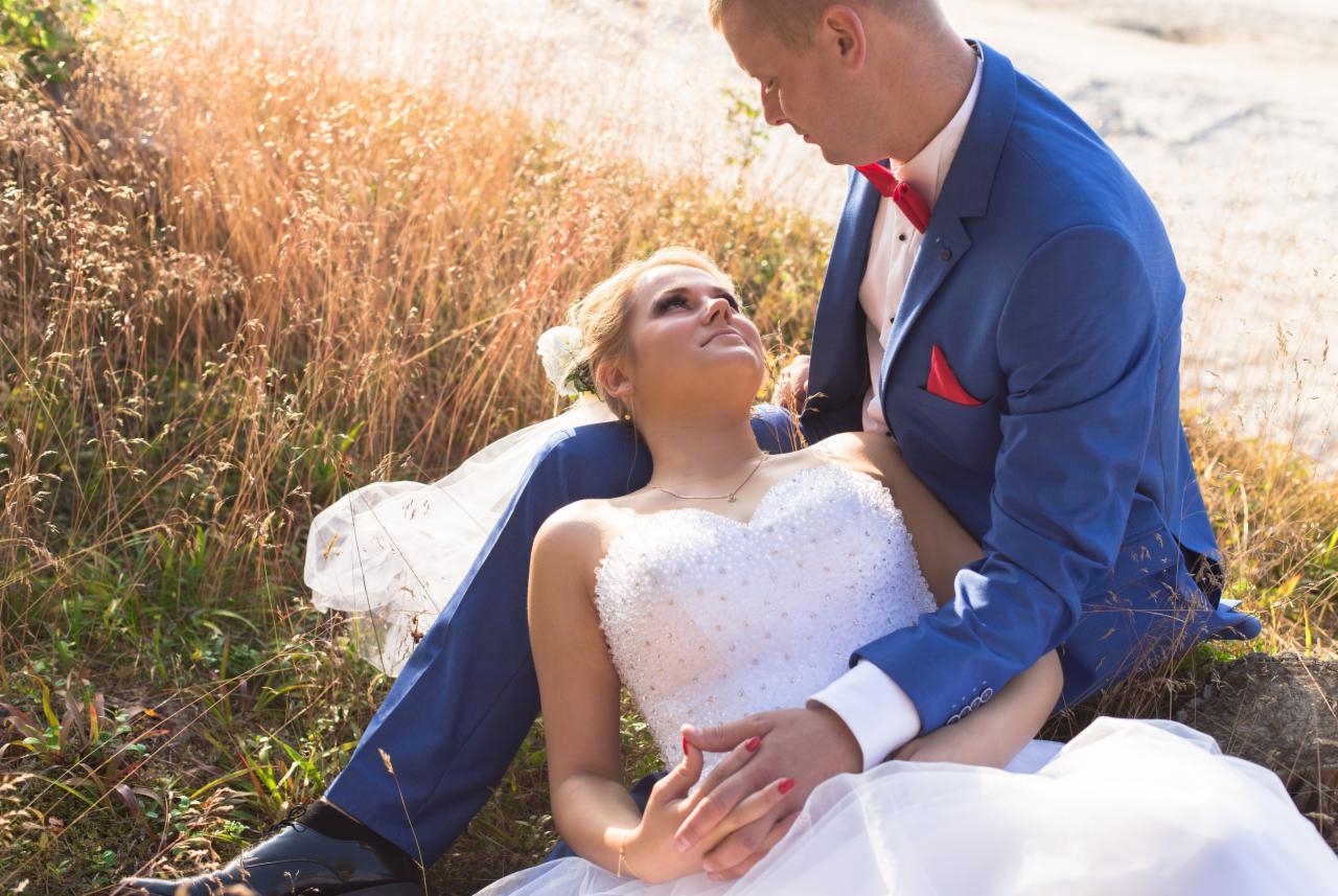 Panna młoda w białej sukni z kryształkami leży na kolanach pana młodego w błękitnym garniturze z czerwoną muchą i poszetką, oboje patrzą na siebie na tle wysokiej trawy.