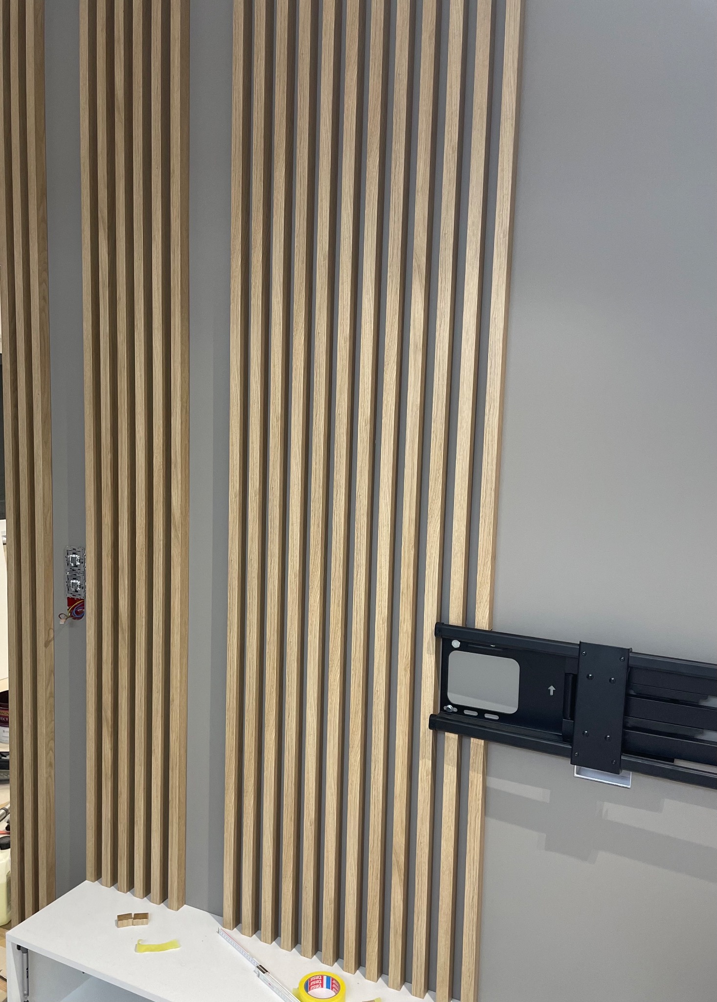 Drewniane lamele na szarej ścianie, z zamontowanym uchwytem do telewizora, widoczne elementy instalacji elektrycznej oraz miarka i taśma klejąca na blacie.
