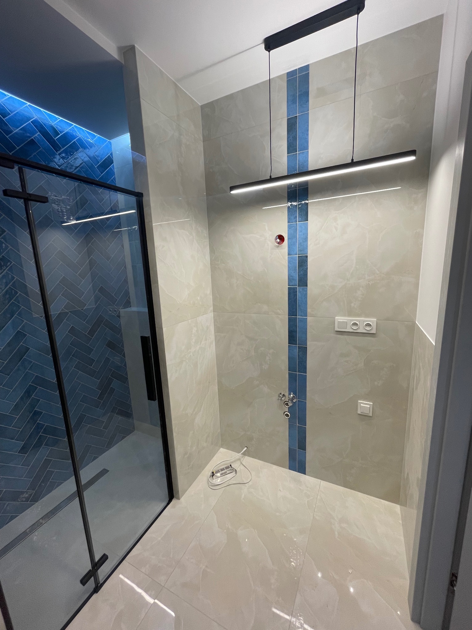 Nowoczesna łazienka z kabiną prysznicową typu walk-in wykończoną niebieską mozaiką w jodełkę, beżowe płytki na ścianach z pionowym pasem niebieskich akcentów, minimalistyczna lampa sufitowa...