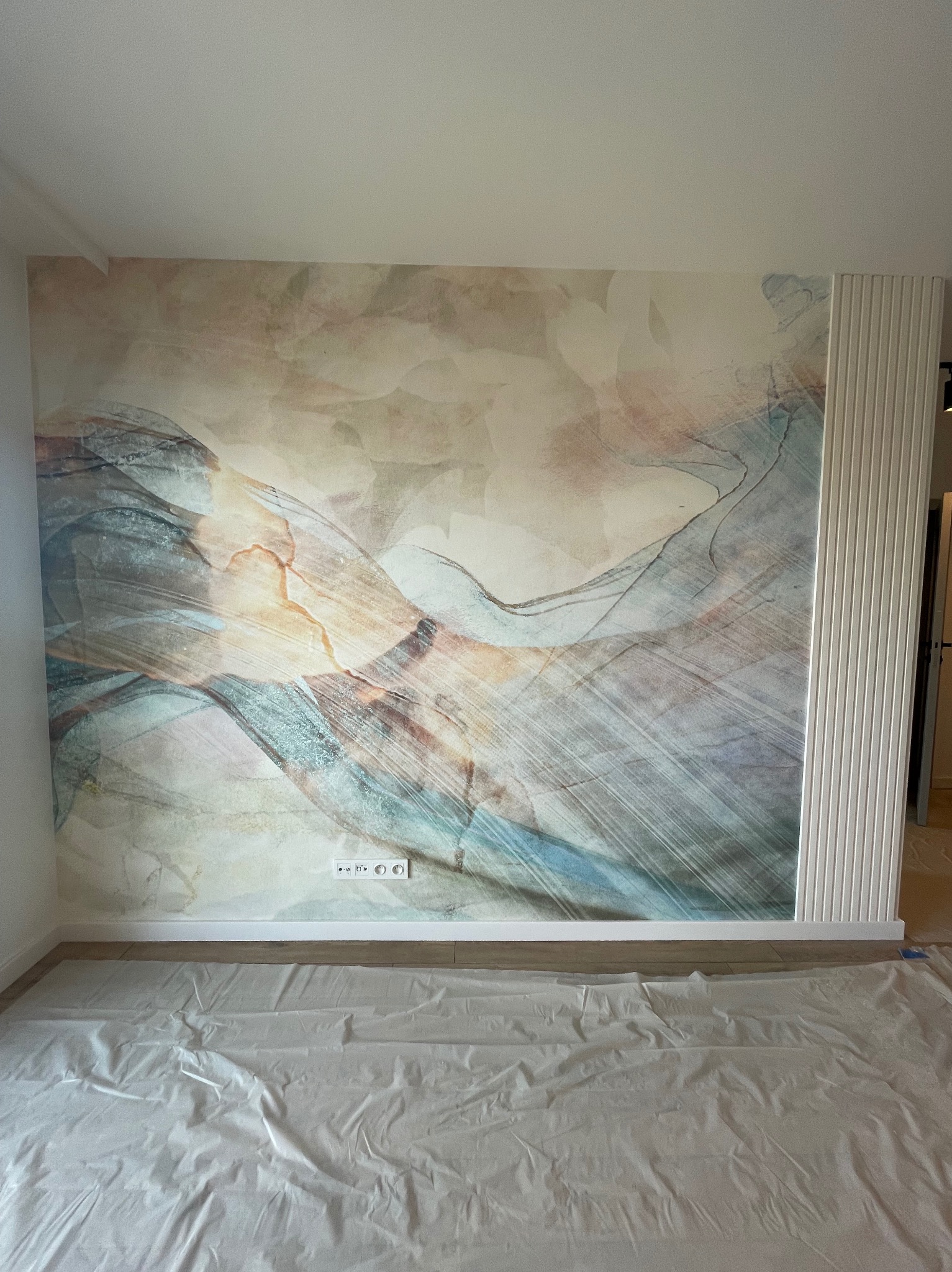 Abstrakcyjna fototapeta w odcieniach beżu, błękitu i szarości na ścianie, zabezpieczona podłoga folią malarską, biała listwa dekoracyjna z pionowymi żłobieniami po prawej stronie.