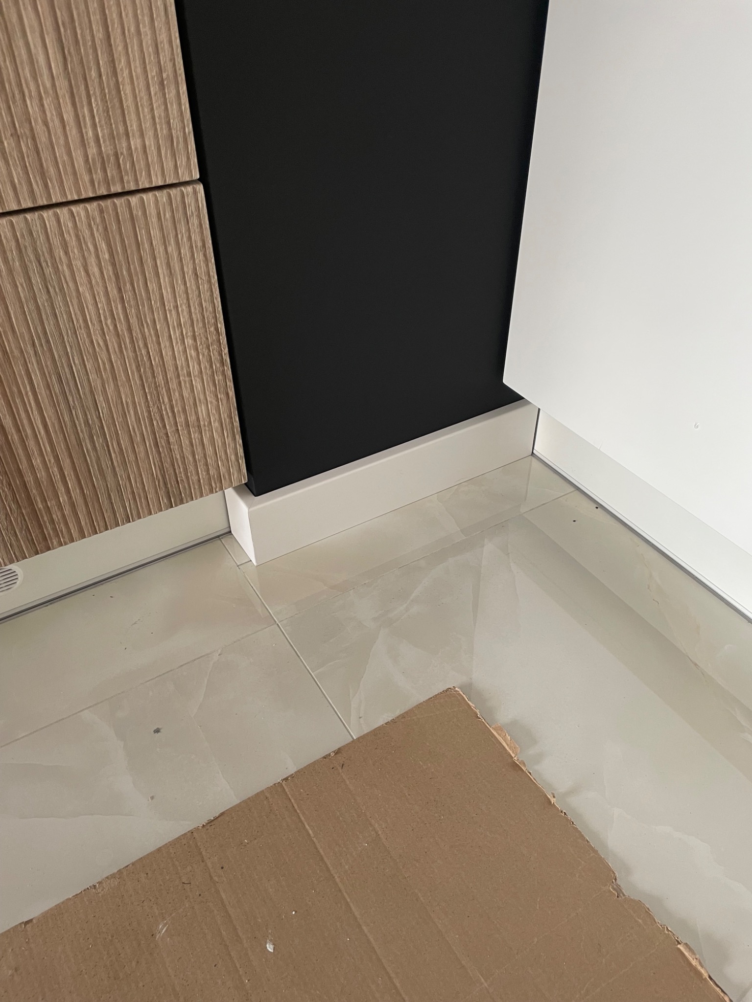 Fragment zabudowy kuchennej z ryflowanym frontem, czarną ścianą i białą szafką, widoczne białe listwy przypodłogowe i błyszczące płytki podłogowe zabezpieczone kartonem.