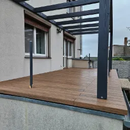 Nowoczesny taras z desek kompozytowych w kolorze drewna, z elementami pergoli w kolorze grafitowym, połączony z budynkiem o jasnej elewacji, widok z boku z fragmentem rynny i wózkiem z drewnem w tle.