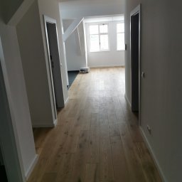 Jakobbauservice Innenausbau - Korytarz z nową podłogą z desek w odcieniach brązu, widoczne białe ściany, drzwi i listwy przypodłogowe, w tle jasne pomieszczenie z oknami.