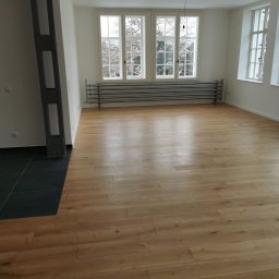 Jakobbauservice Innenausbau - Jasna drewniana podłoga ułożona w nowym, przestronnym pokoju z dużymi oknami i ciemnymi płytkami przy metalowym słupie konstrukcyjnym.