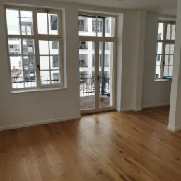 Jasny, przestronny pokój z drewnianą podłogą ułożoną w deski, białymi ścianami z trzema oknami z widokiem na balkony sąsiedniego budynku w zimowej scenerii.