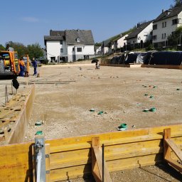 Jakobbauservice Innenausbau - Przygotowanie terenu pod fundamenty domu: drewniane szalunki, wyrównany grunt z wystającymi rurami instalacyjnymi, w tle ekipa budowlana przy pomarańczowym samochodzie.