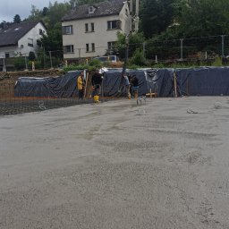 Jakobbauservice Innenausbau - Wylewanie betonu na przygotowany teren zbrojony siatką, widoczni pracownicy w żółtych gumowcach i maszyna do pompowania betonu, w tle zabudowa jednorodzinna i zieleń.
