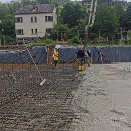 Jakobbauservice Innenausbau - Wylewanie betonu na siatkę zbrojeniową; widoczna pompa do betonu, ekipa w żółtych gumowcach oraz budynek mieszkalny w tle.