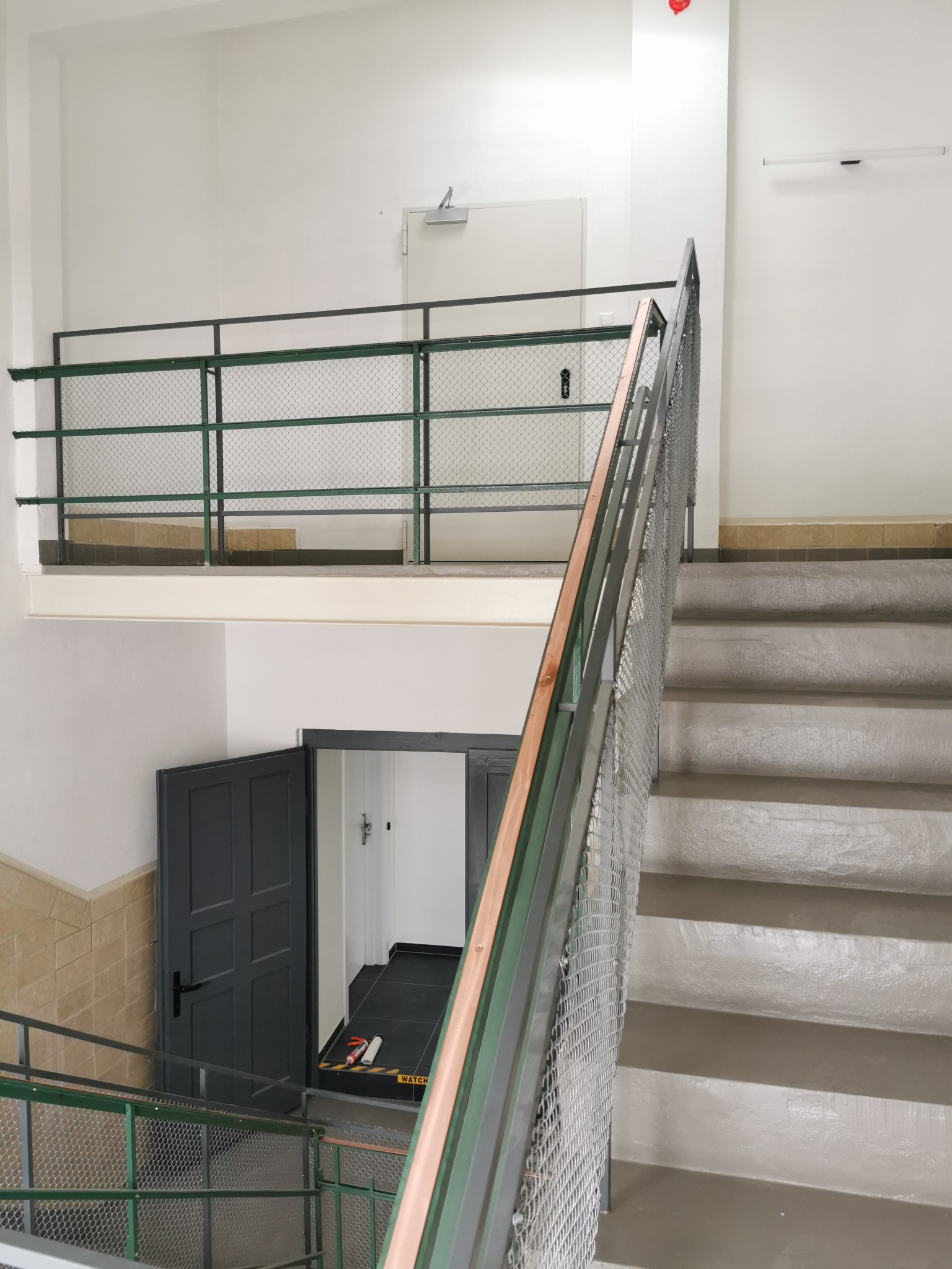 Klatka schodowa po remoncie, z szarymi schodami, metalową siatką zabezpieczającą i pomalowanymi balustradami, widoczne drzwi na półpiętrze i fragment korytarza z czarną podłogą i żółtymi taśmami...