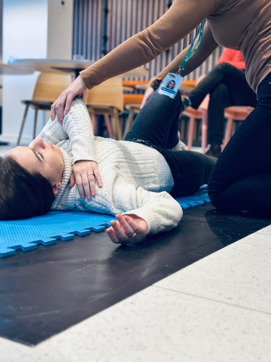 Instruktor z identyfikatorem na smyczy delikatnie stabilizuje ramię kobiety leżącej na niebieskiej macie podczas ćwiczenia.
