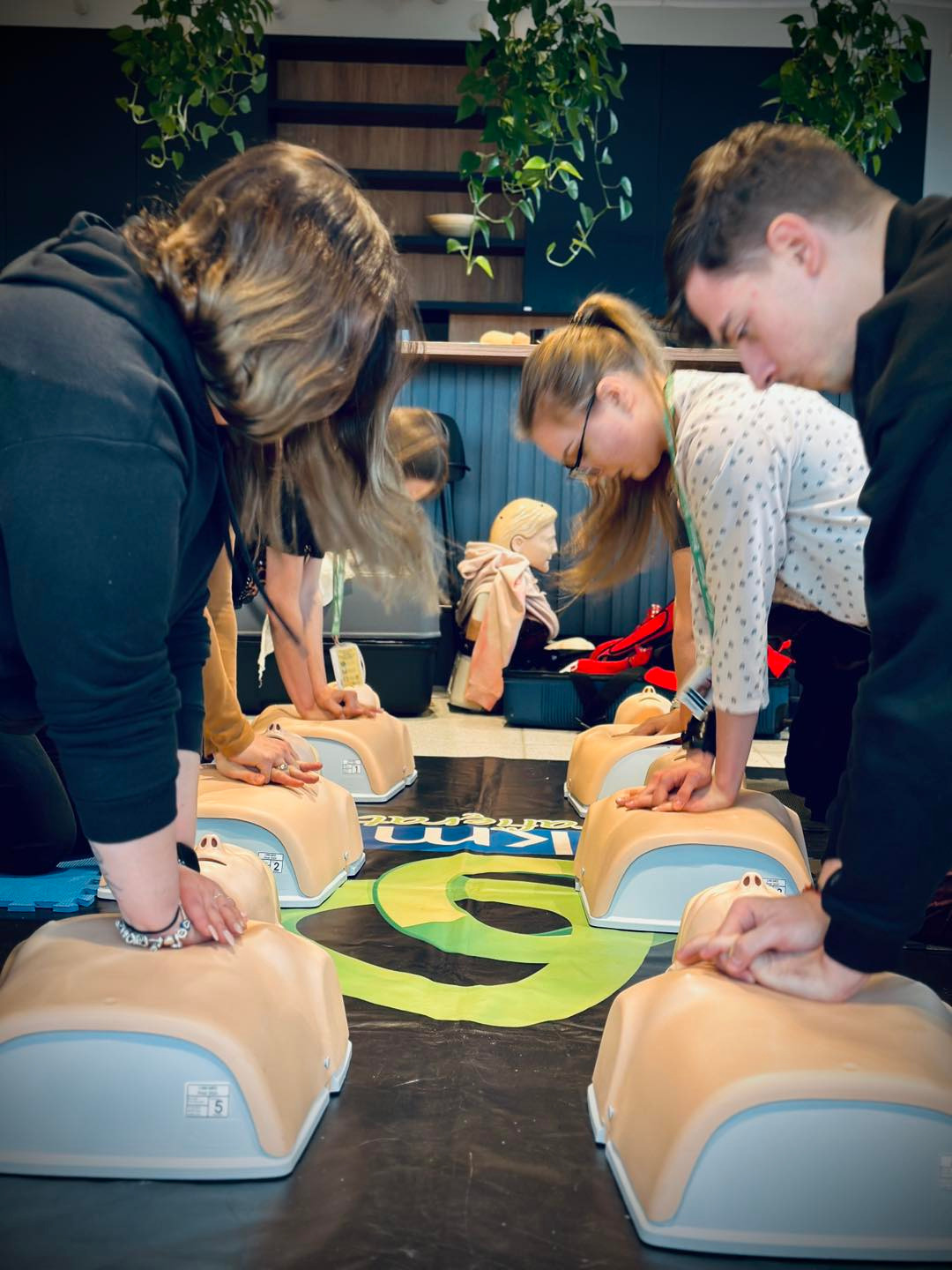 Grupa osób ćwiczy resuscytację krążeniowo-oddechową na fantomach na czarnej macie z zielonymi wzorami w pomieszczeniu z roślinami.