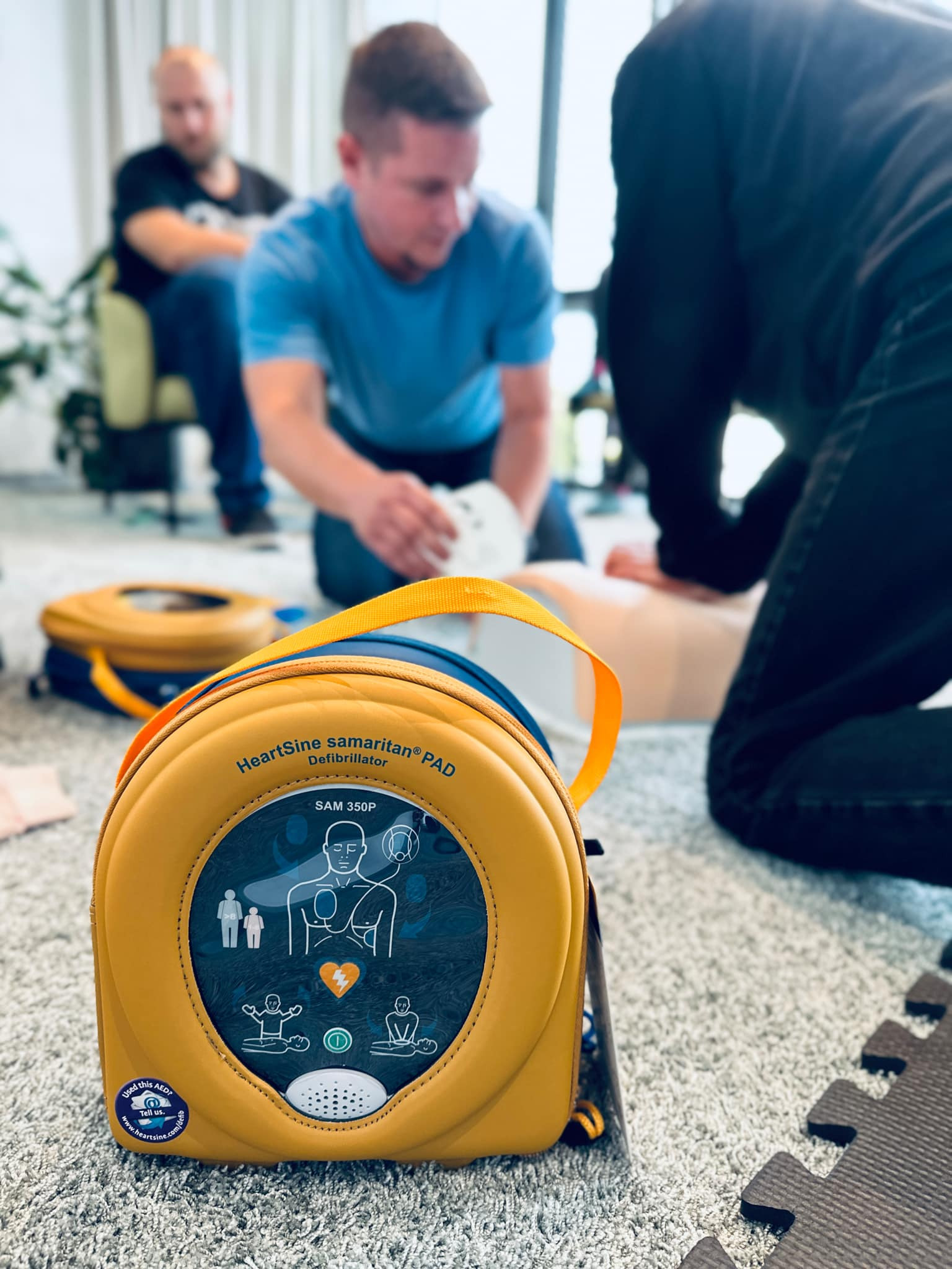 Żółto-niebieska torba defibrylatora HeartSine Samaritan PAD SAM 350P na dywanie, w tle instruktorzy ćwiczący resuscytację na manekinie.