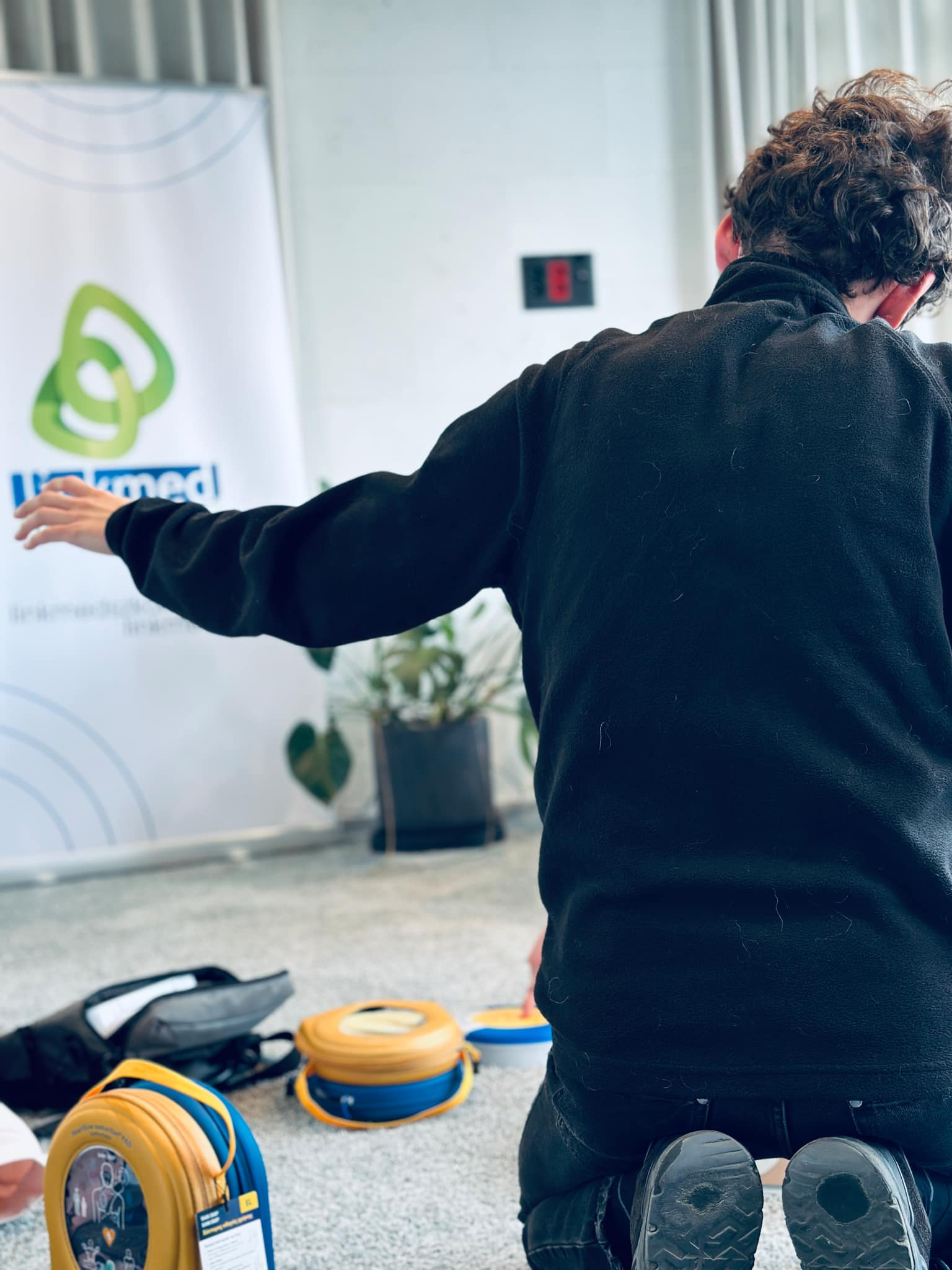 Osoba klęczy na podłodze, wyciągając rękę w stronę plakatu z logo firmy, w pobliżu leżą torby i defibrylatory treningowe.