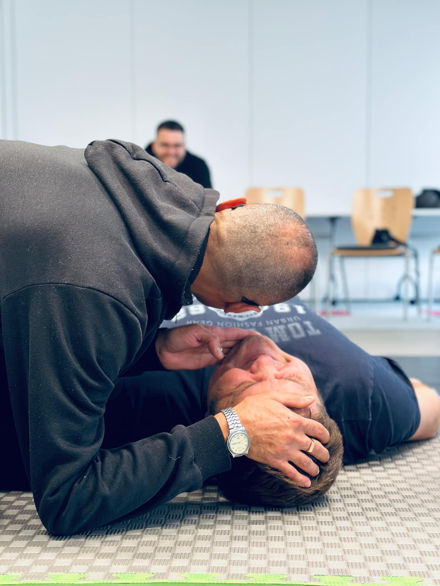 Instruktor w czarnej bluzie demonstruje na leżącym mężczyźnie z zegarkiem na ręce, jak udrożnić drogi oddechowe; w tle obserwator.