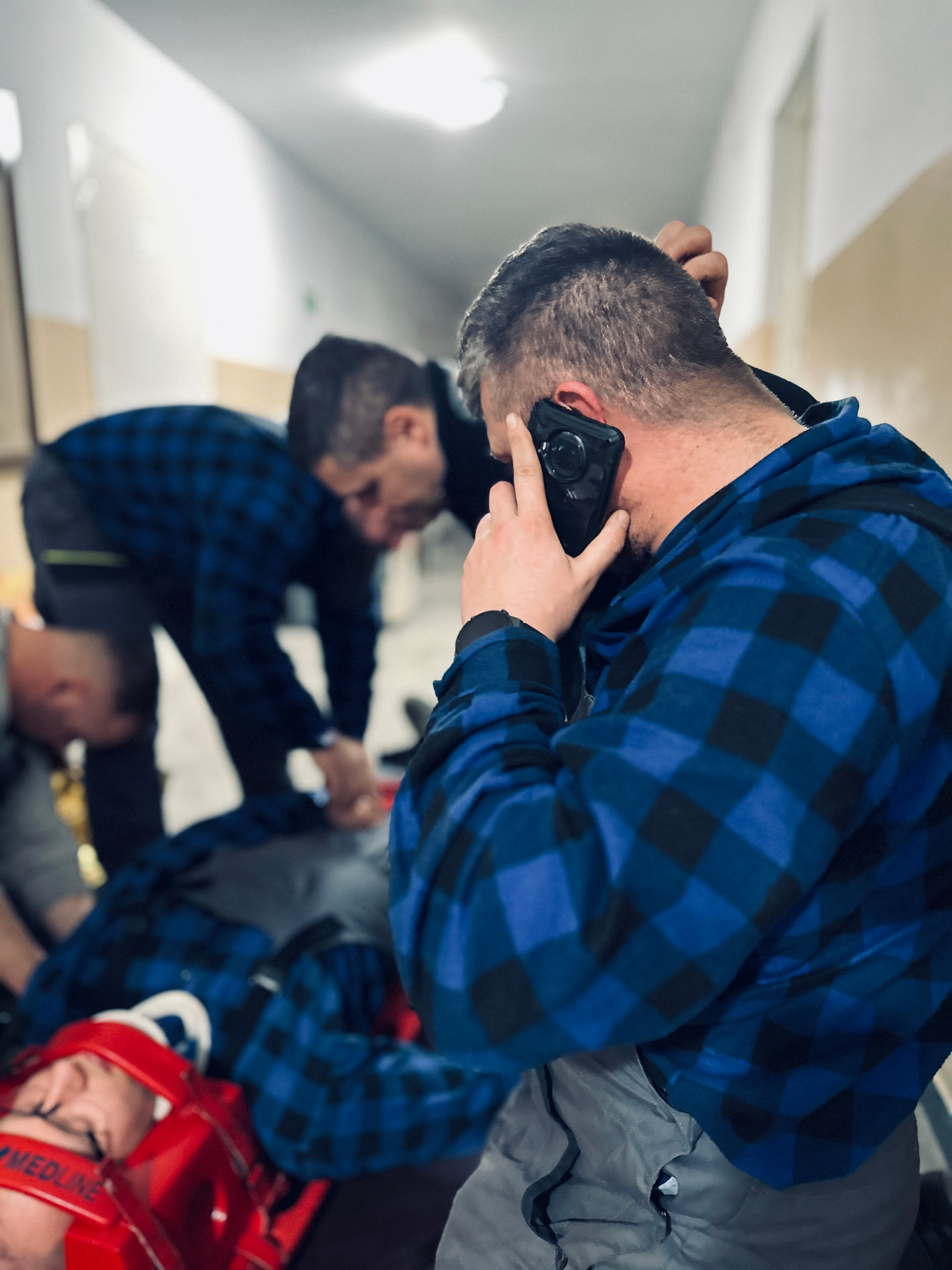 Instruktor rozmawia przez telefon podczas symulacji udzielania pomocy poszkodowanemu leżącemu na noszach z stabilizacją głowy, w tle inni uczestnicy szkolenia.
