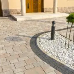 Jasnoszara kostka brukowa ułożona wzorem cegły, obrzeże z ciemniejszych kostek, lampa ogrodowa i ozdobny krzew na białych kamieniach przed wejściem do domu z beżową elewacją.