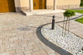 Jasnoszara kostka brukowa ułożona wzorem cegły, obrzeże z ciemniejszych kostek, lampa ogrodowa i ozdobny krzew na białych kamieniach przed wejściem do domu z beżową elewacją.