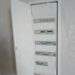 Otwarta, biała skrzynka rozdzielcza z licznikami i bezpiecznikami na ścianie w Kłobucku, widoczne numery od 1 do 60.