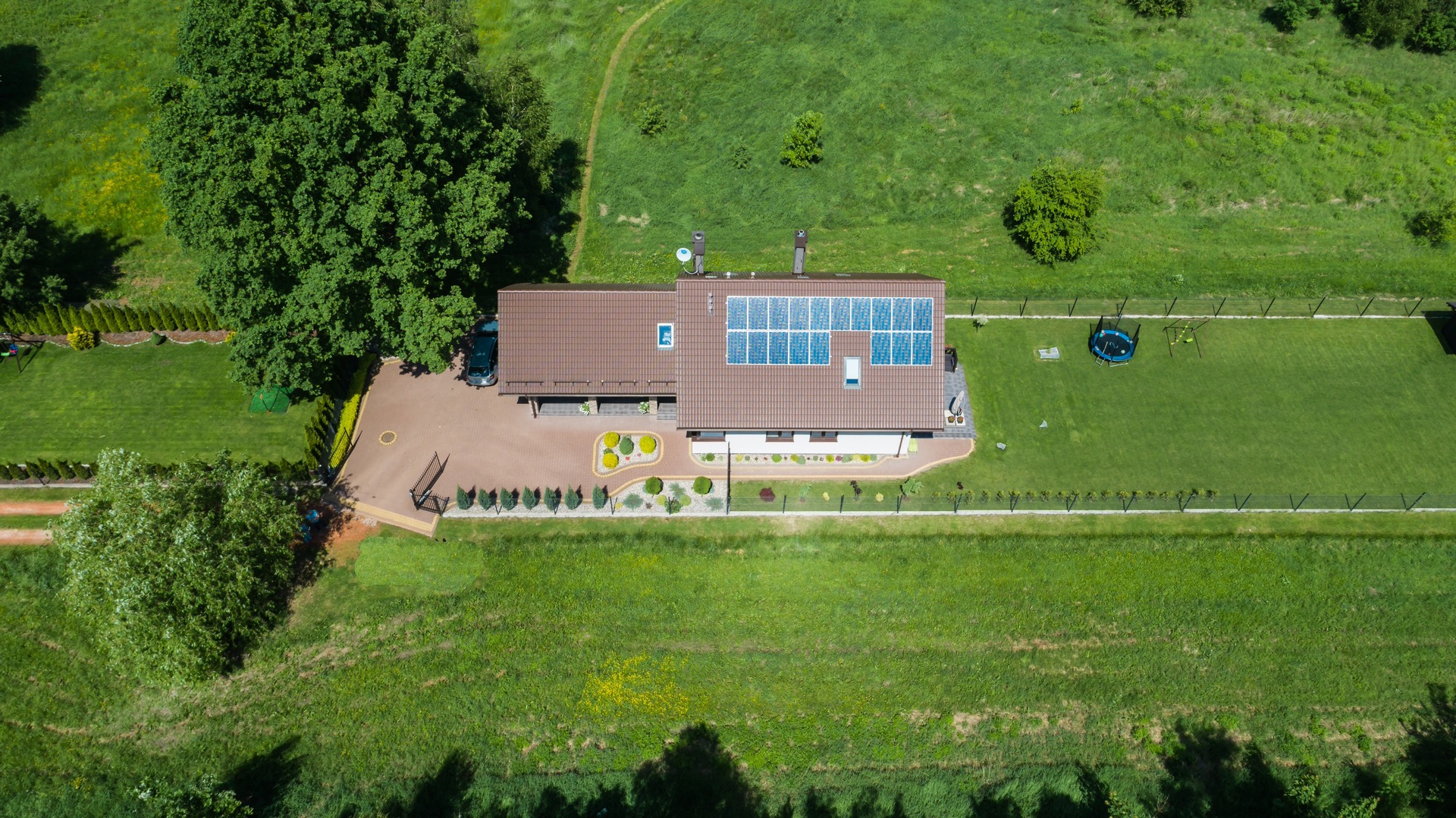 Widok z drona na dom jednorodzinny z brązowym dachem i panelami fotowoltaicznymi na dachu, otoczony zadbanym ogrodem z trampoliną i miejscem parkingowym dla samochodu.