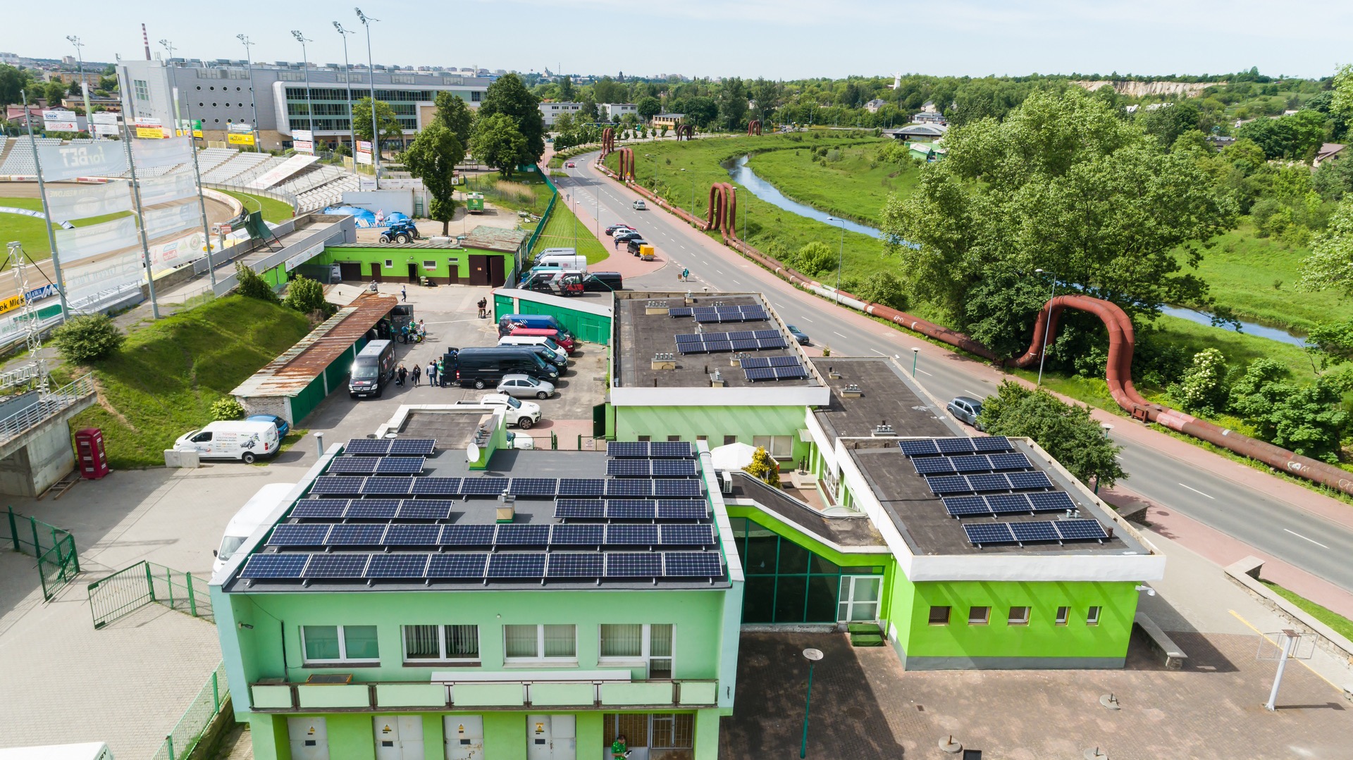 Widok z góry na budynki z panelami słonecznymi na dachu, obok stadion, rzeka i charakterystyczne rury ciepłownicze w tle.