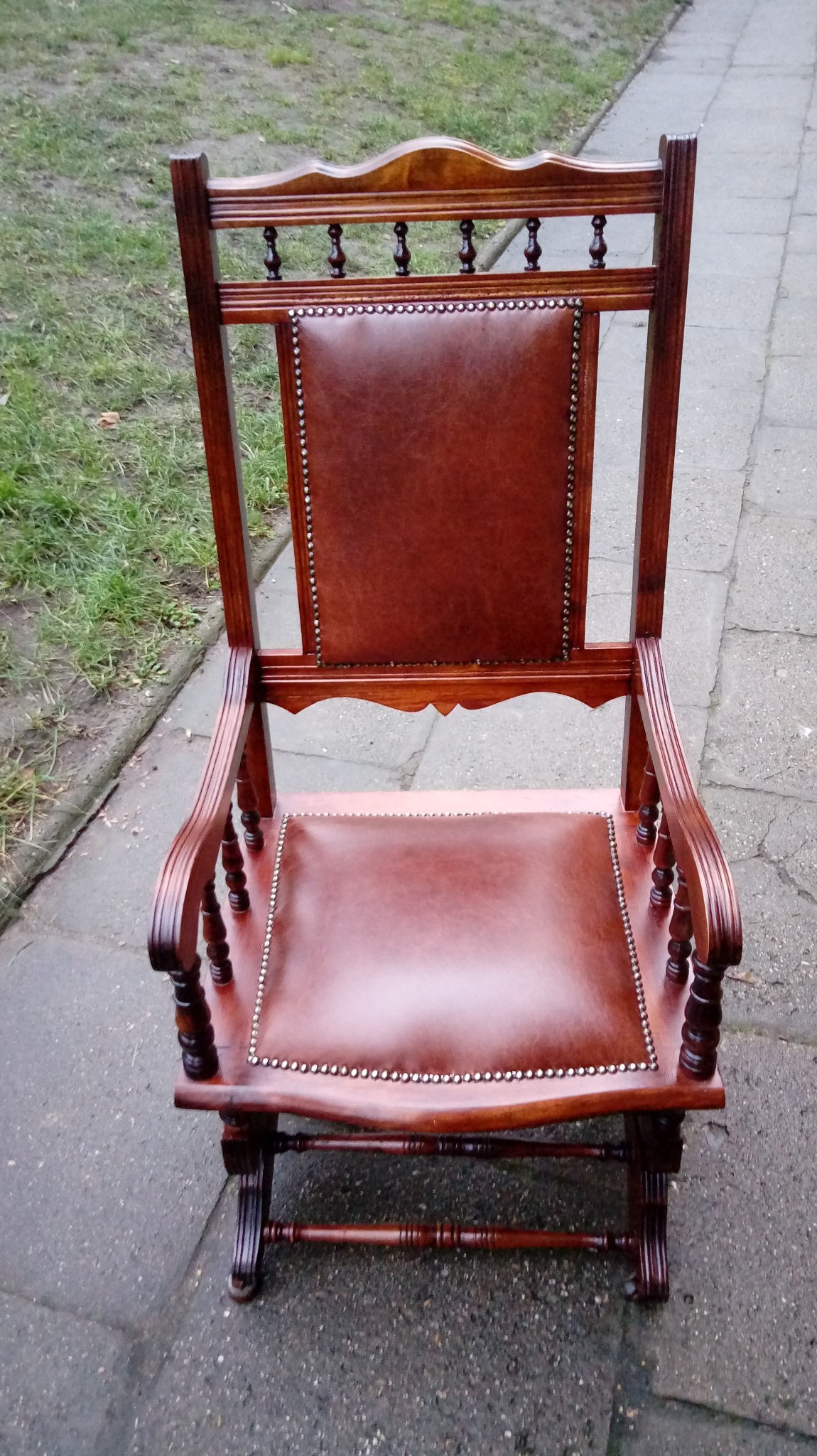 Odnowiony fotel bujany z brązową skórzaną tapicerką, ozdobiony ćwiekami, stojący na chodniku obok trawnika.