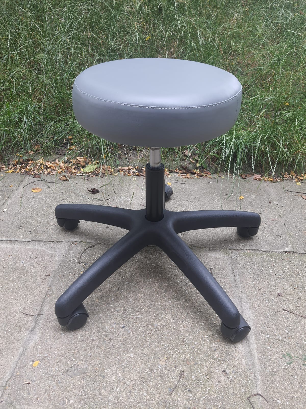 Szary taboret obrotowy z tapicerowanym siedziskiem na czarnej podstawie z kółkami, stojący na betonowej powierzchni z zielonym tłem trawy.