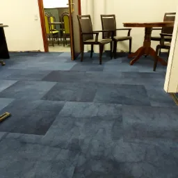 Fragment biura z ciemnoniebieską wykładziną w kwadraty, widoczny odkurzacz, krzesła i stół w tle